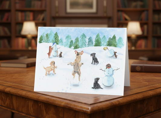A folded holiday card stands upright on a wooden desk. The front features a hand-painted winter scene with several playful puppies and dogs in the snow, all in front of a background of evergreen trees and a softly painted winter sky. One puppy jumps into the air joyfully in the center, while others play, sit, or help build snowmen nearby. The card has a soft, whimsical watercolor style.