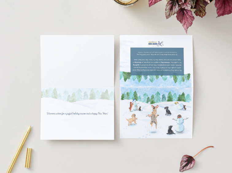 Two cards are shown on a light tabletop:

The left card is opened, revealing an interior design with a pale snowy landscape across the middle and the printed message: “Warmest wishes for a joyful holiday season and a happy New Year!”
The right card is folded flat and placed face-up. Its front displays the snowy puppy scene, while the back shows the Guide Dog Foundation logo, a blue information panel with text, and the continuation of the snowy painted landscape. 