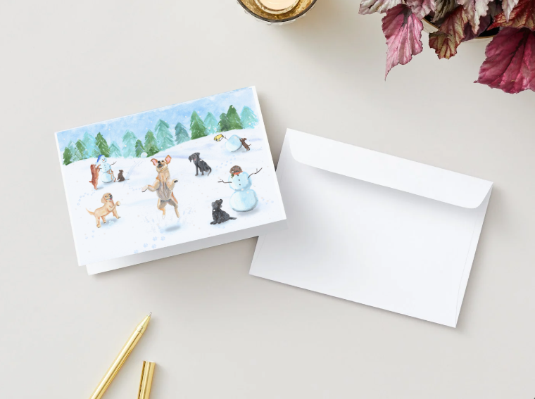 A holiday card is shown partially folded on a tabletop next to a plain white envelope. The card’s cover displays the illustrated winter scene of puppies playing in the snow—some building snowmen, some sitting, and one leaping excitedly in the center. The watercolor pine trees and sky create a soft, festive backdrop. 